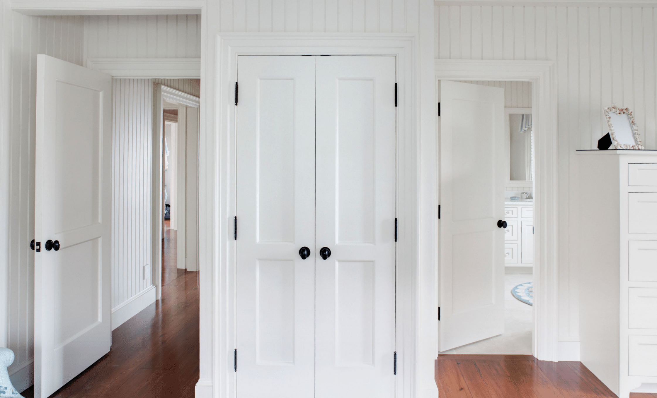 A hallway with wooden floors features three open white doors and a closed double-door closet. The walls are white with vertical paneling, and a glimpse of a bathroom and another room is visible in the background.