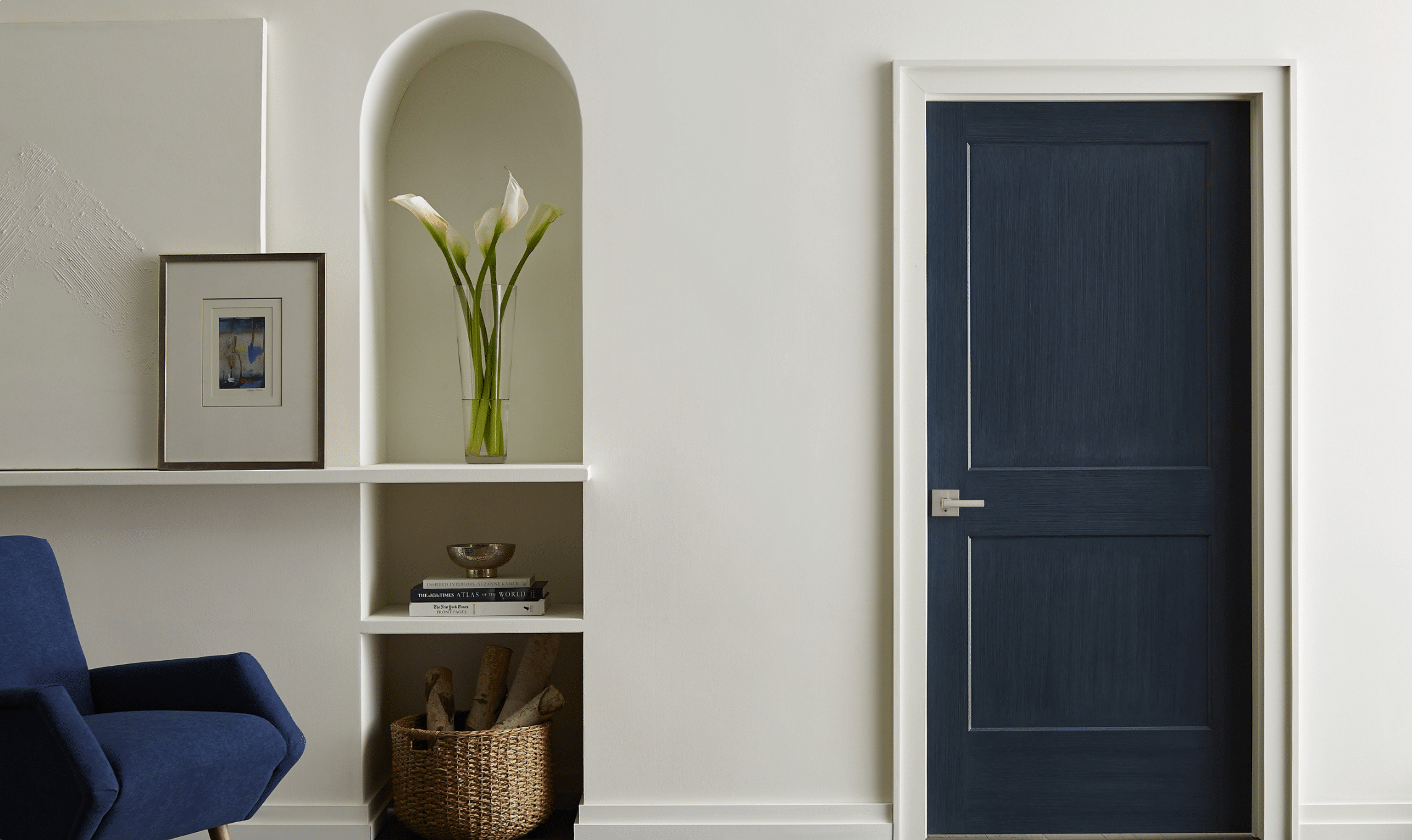 A modern interior with a dark blue door, a blue armchair, white walls, a built-in shelf holding artwork, a basket of firewood, and a vase with calla lilies in a small arched alcove.