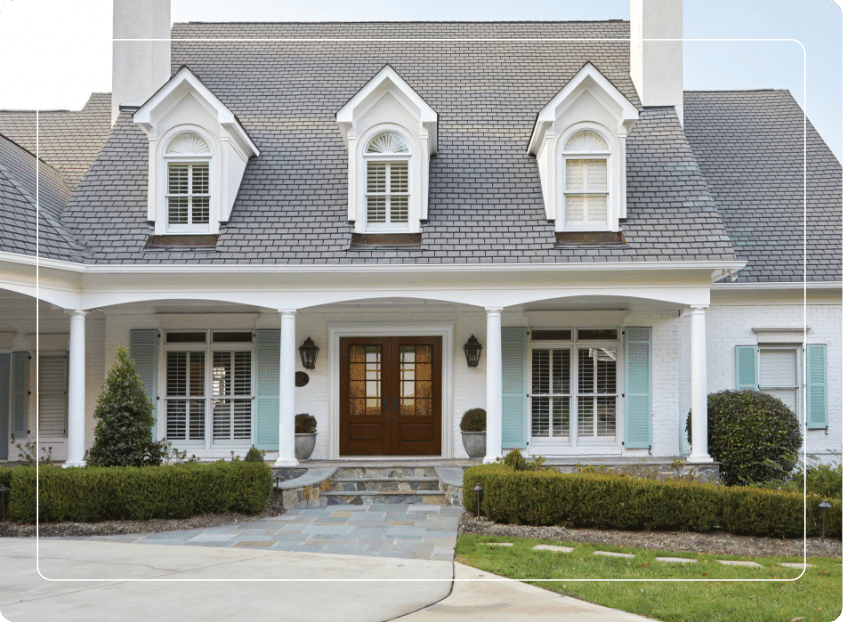 A two-story house with a gray shingle roof, white brick exterior, blue shutters, and a wooden double front door. The house features three dormer windows and a covered front porch with white columns.