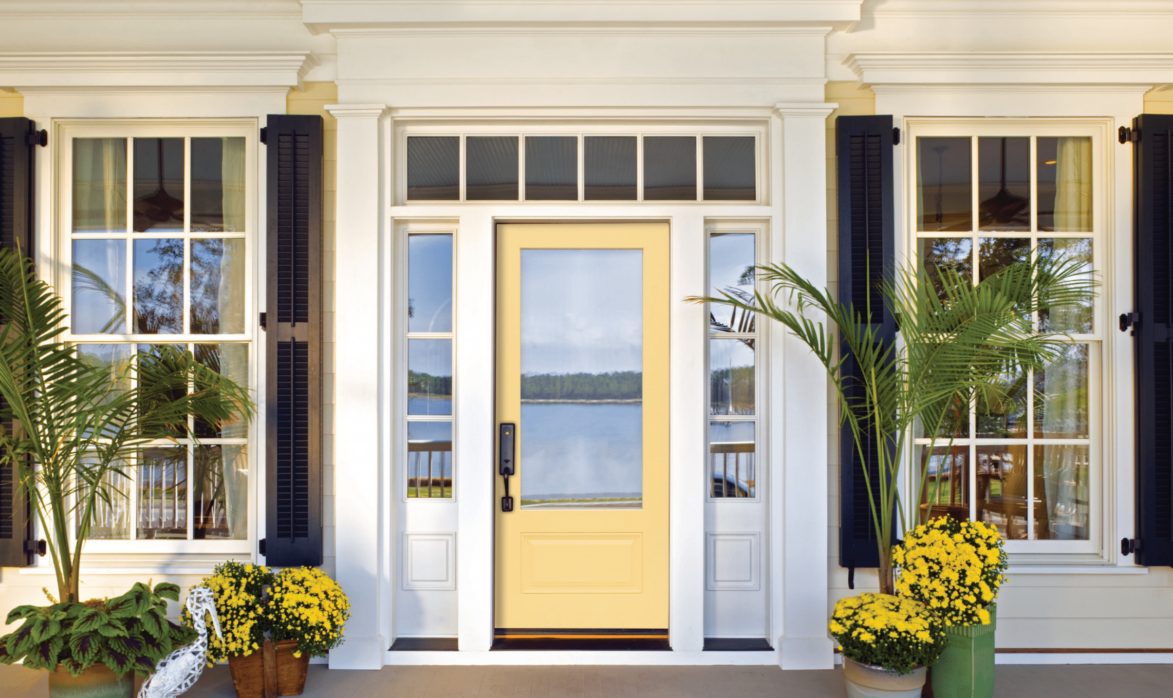 A yellow front door with glass panels is framed by white trim and flanked by tall windows with black shutters. Potted palms and yellow flowers decorate the porch, with a lake and trees visible through the doorway.