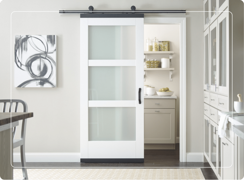 A modern kitchen with a white sliding barn door partially open, revealing a neatly organized pantry with shelves, jars, and green apples on the counter. Abstract art hangs on the adjacent wall.