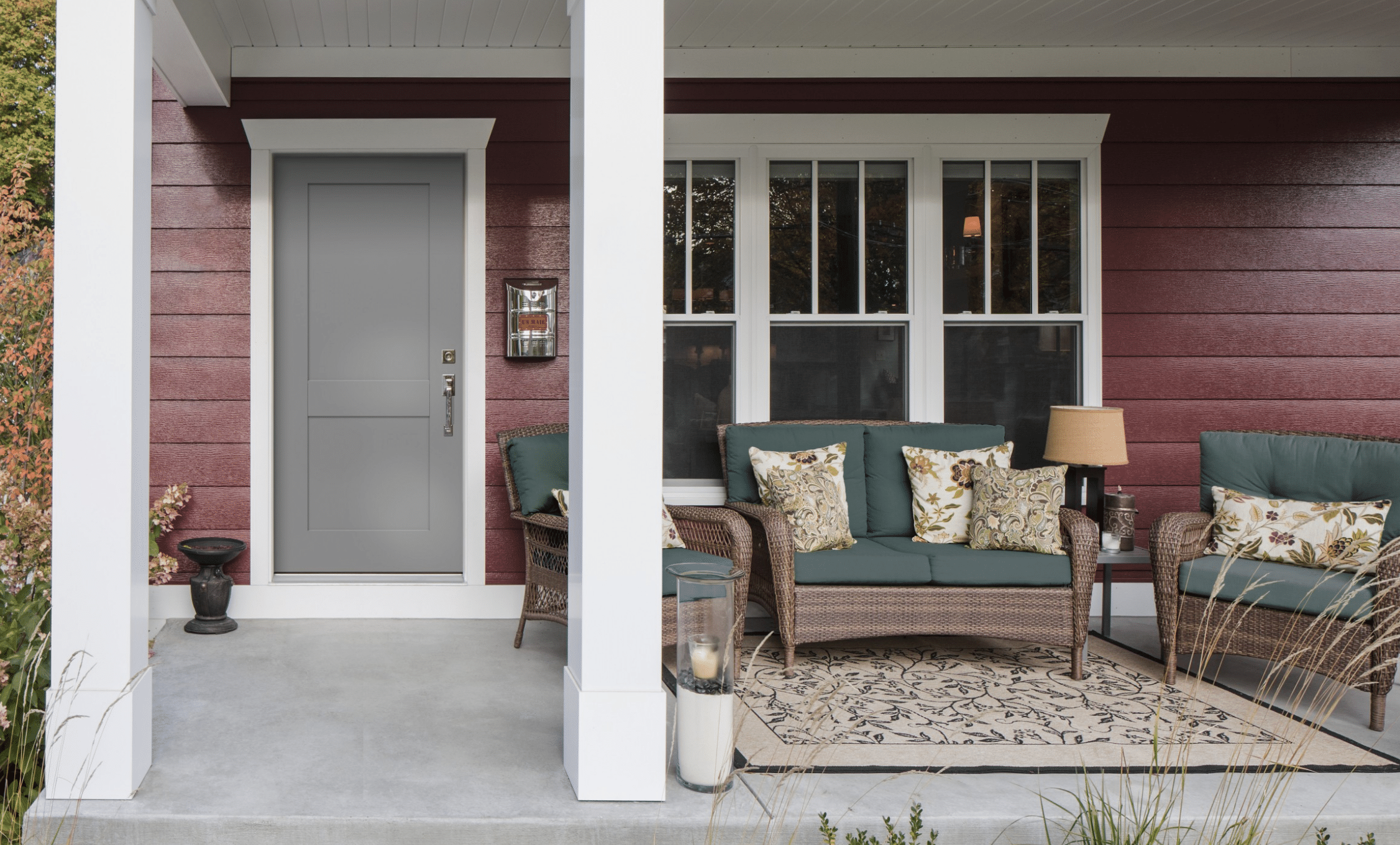 A cozy front porch with a red exterior, gray door, and white trim. There is wicker seating with green cushions and floral pillows, a small table with a lamp, and a patterned rug on the concrete floor.
