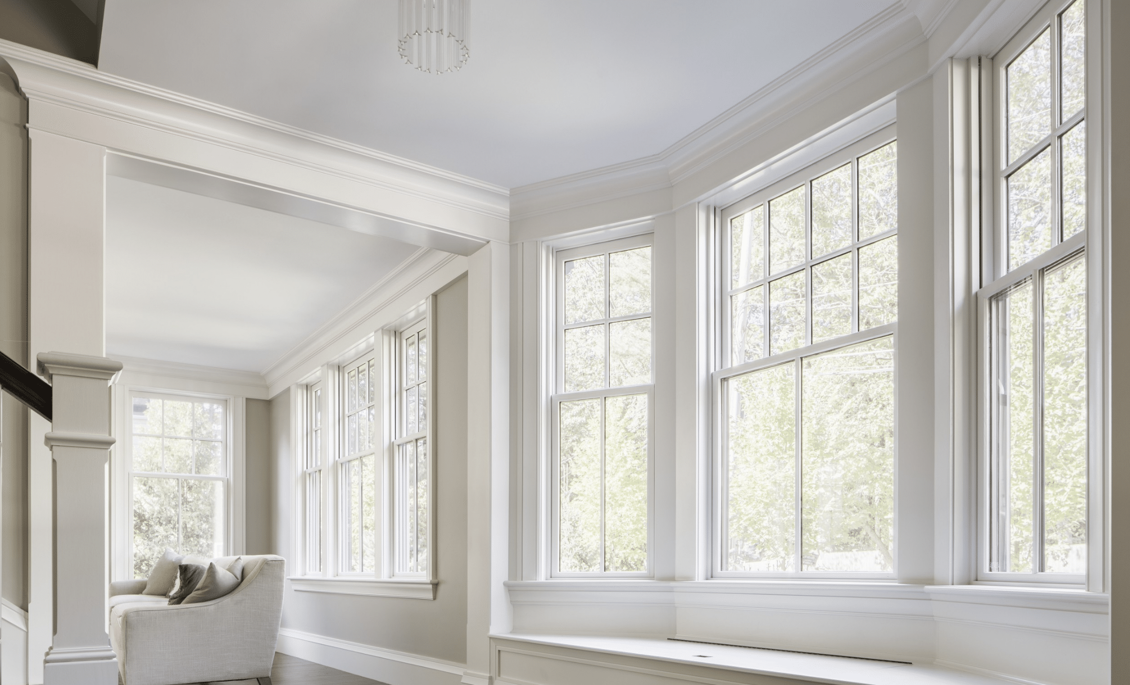 Bright sunroom with large bay windows, white trim, and built-in bench seating. A light-colored armchair sits in the corner, and trees can be seen through the windows. The room has a clean, airy feel.