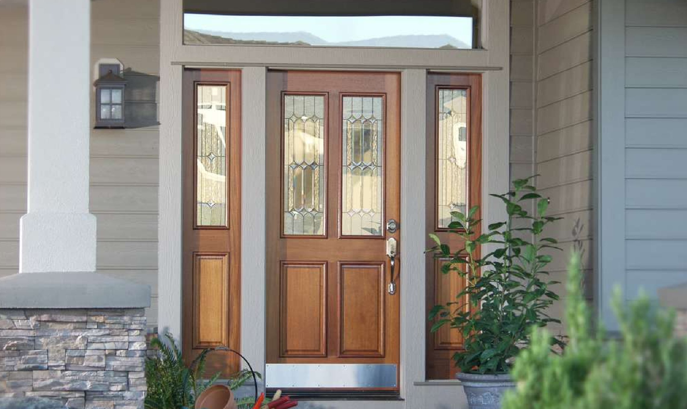 A wooden front door with decorative glass panels is framed by two glass sidelights. The entryway features stone accents and potted plants on either side.